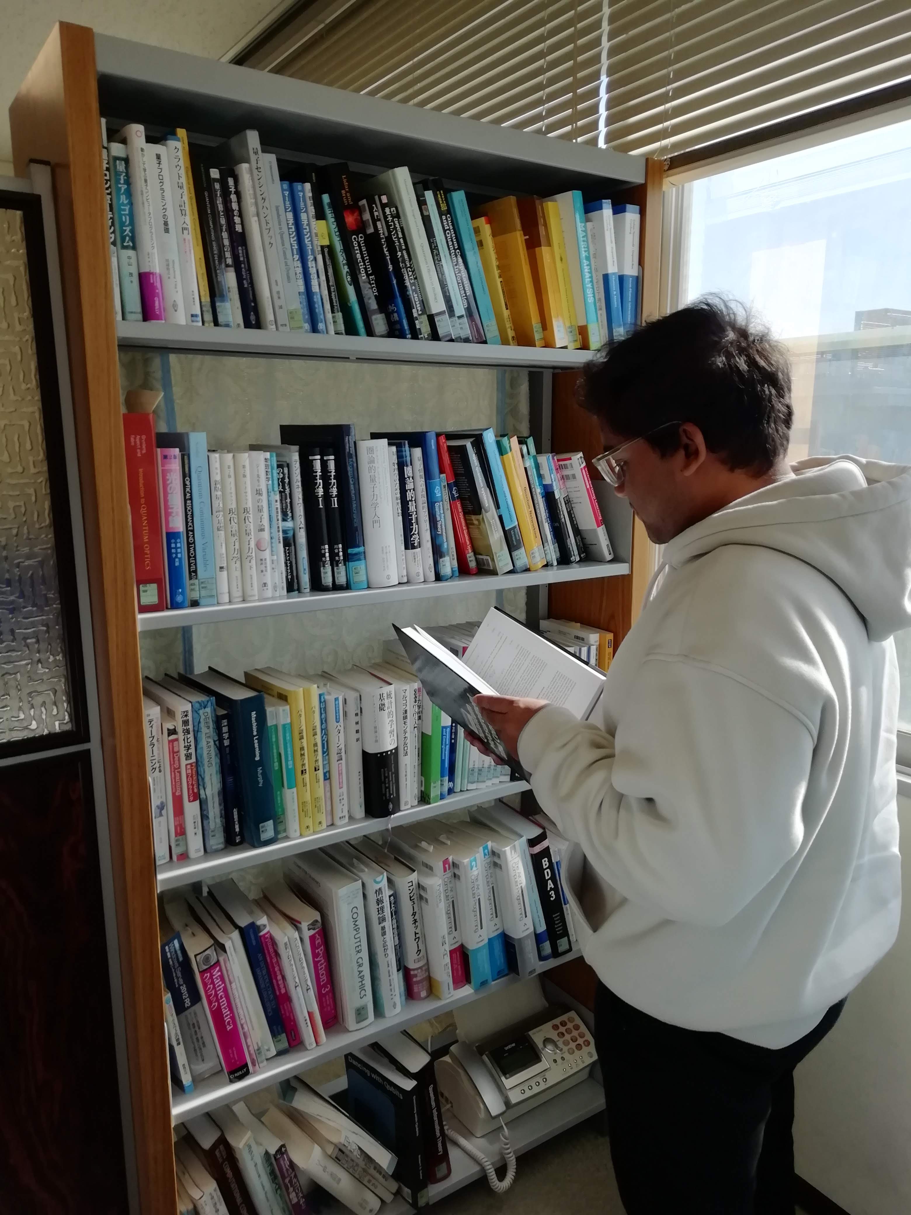 研究室の本棚から学術書を取り出し読むインドからの留学生。理論やシミュレーションを中心とした研究を進めるためには、様々な数学の定理や情報科学の技術を用いる必要があります。それらの定理や技術を学ぶために、研究室には多数の学術書を揃えています。