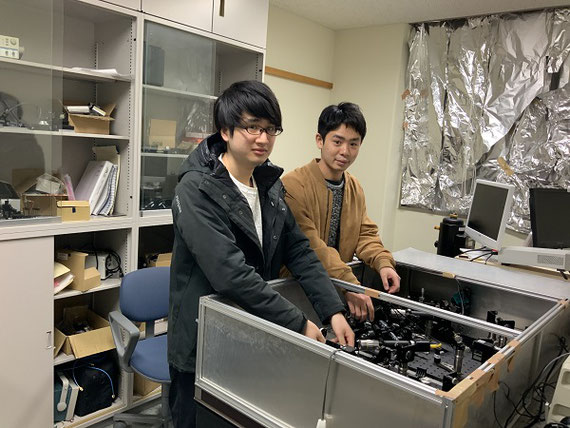 研究風景(レーザー計測)。金属ナノ粒子の性能は、レーザー光を使って試験します。レーザー光は数多くの鏡やレンズを通過した後に金属ナノ粒子に照射します。金属ナノ粒子より現れるごく微弱な光を検出するため、実際の実験は部屋全体を暗くして行います。こちらに写っている２名は大学院生で、新しい光学系の設計について打ち合わせしているところです。