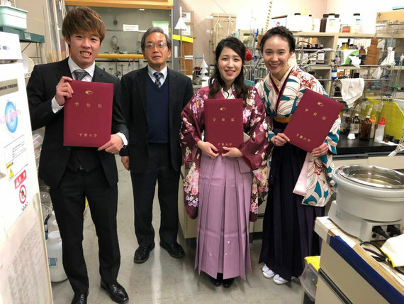 卒業式にて、研究室の卒業生３人と。左が薬品を保管する防爆冷蔵庫、奥がドラフトチャンバー、右にロータリエバポレーターの浴槽のみが写っています。